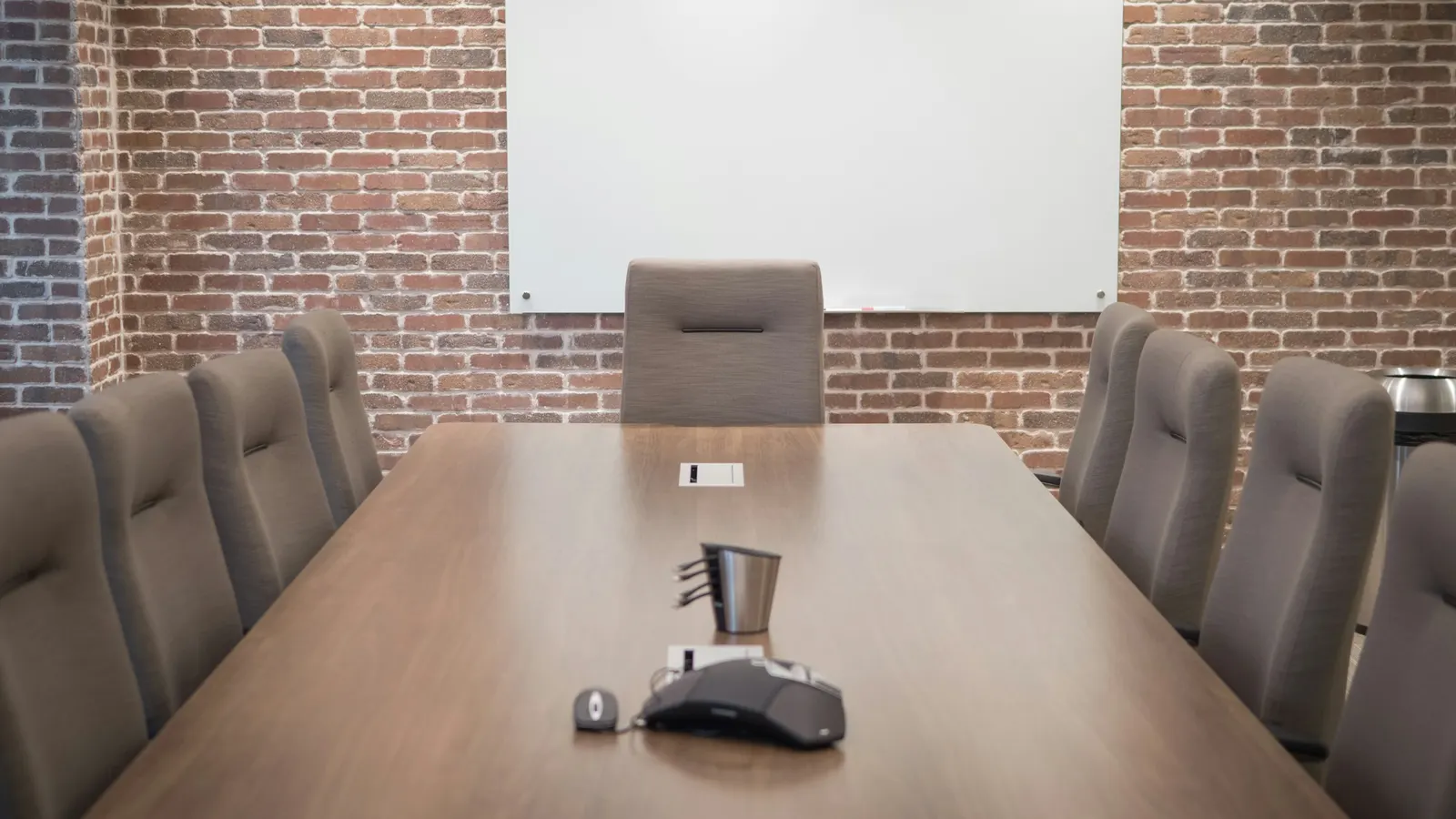 Une salle de réunion vide : une longue table en bois, des fauteuils gris de chaque côté, un mur de briques et un tableau blanc au fond