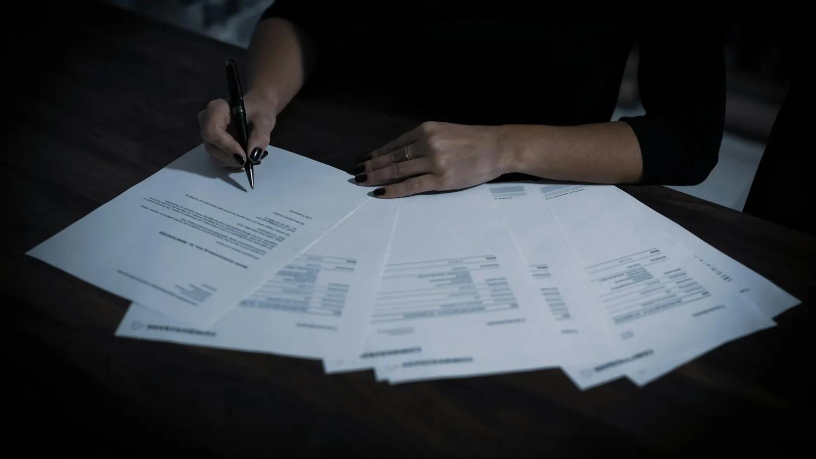 Sur une table sombre, une main tient un stylo au-dessus de plusieurs documents étalés en éventail