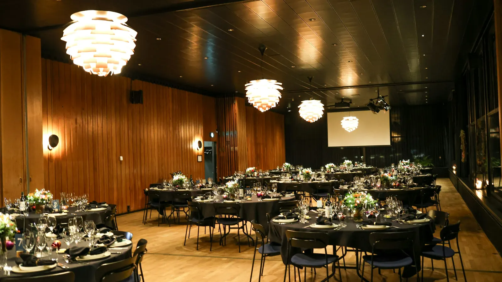 Une salle de réception aux murs de bois, avec des tables rondes dressées sous de grands lustres et un écran de projection au fond