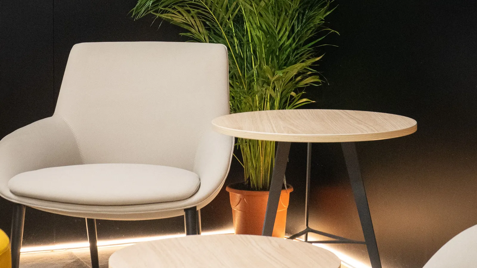 Coin d'un stand équipé : un fauteuil clair, des tables rondes en bois et un palmier en pot devant une cloison noire, sous un liseré lumineux