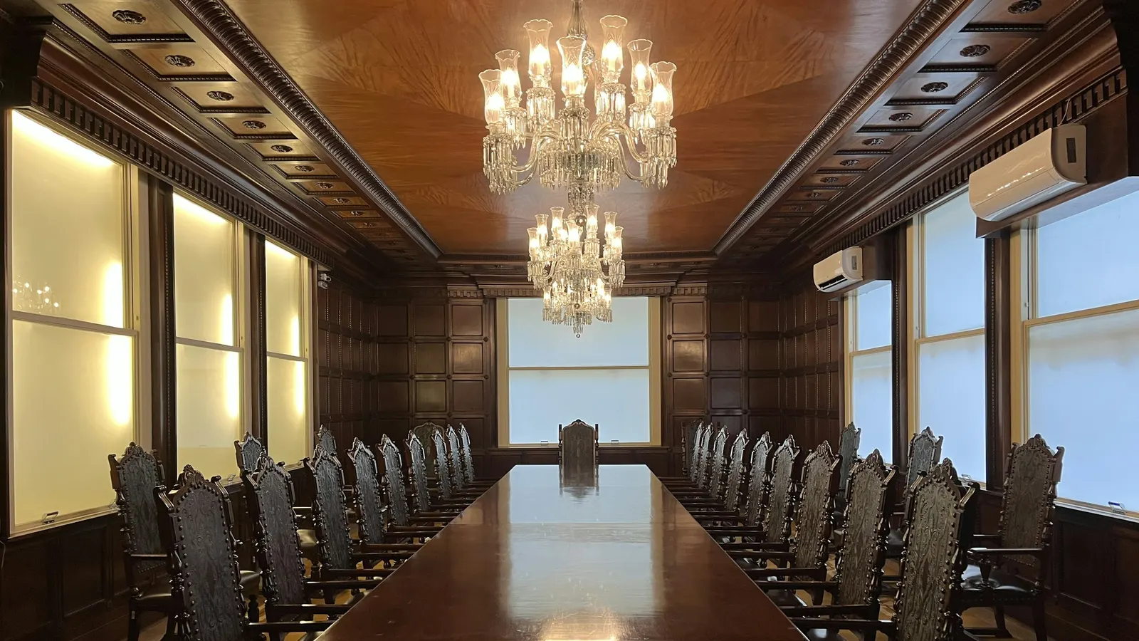Une longue table de réunion en bois verni, bordée de fauteuils sculptés, sous deux lustres de cristal