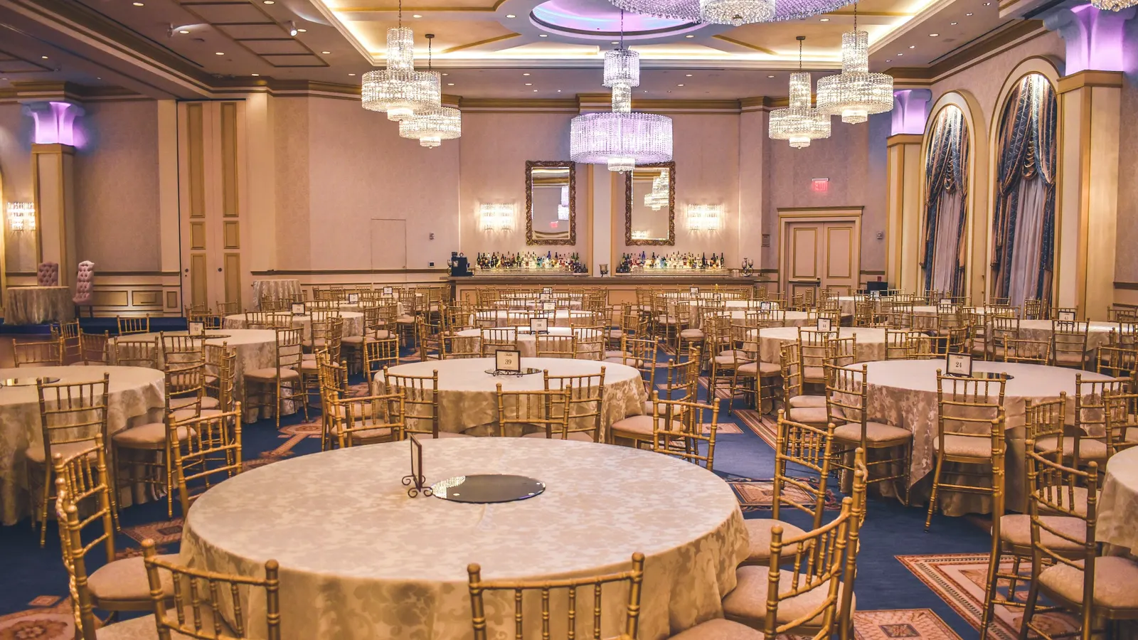 Grande salle de réception : tables rondes aux nappes crème, chaises dorées et lustres de cristal, avec un bar au fond
