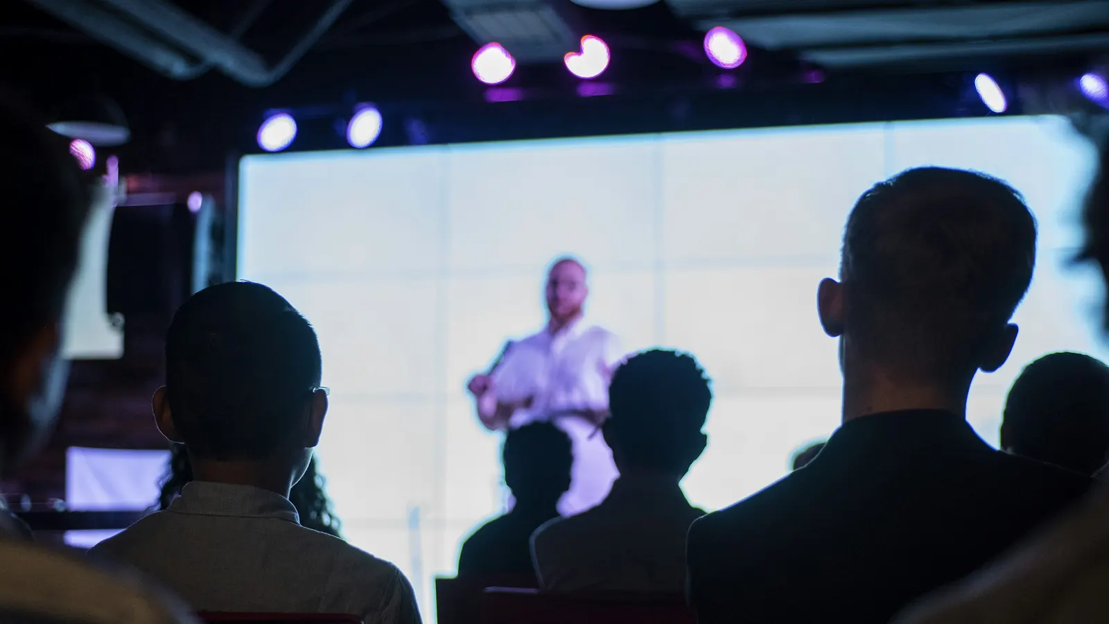 Audience seen from behind in a dark hall, watching a speaker on a large lit screen under stage lights