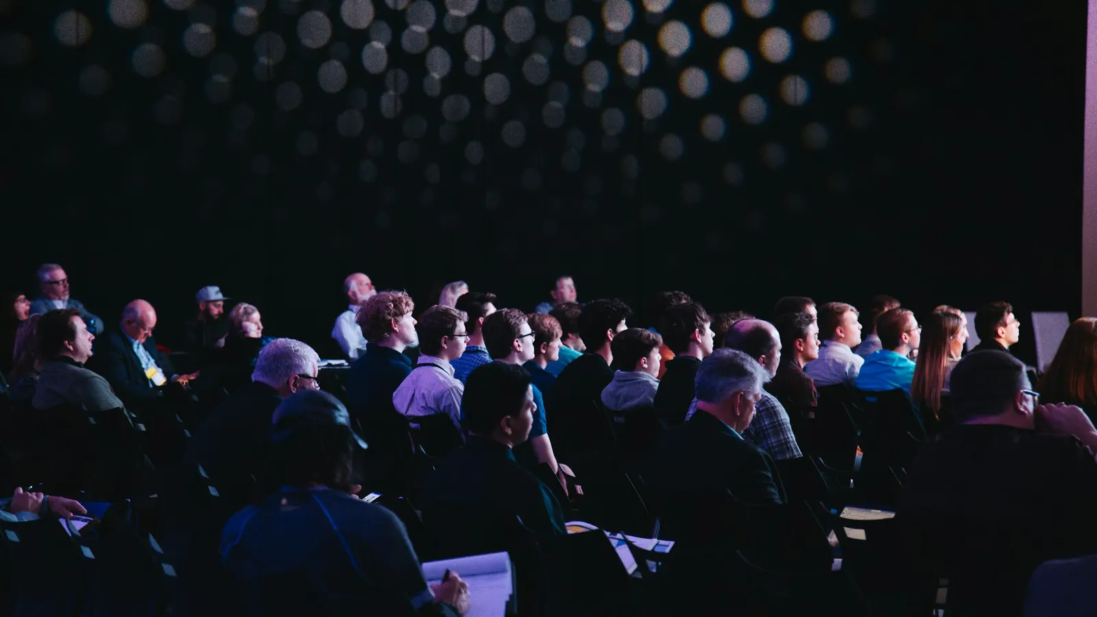 An audience seated in a dark hall at a business event, seen from the side, most of them looking to the right of the frame