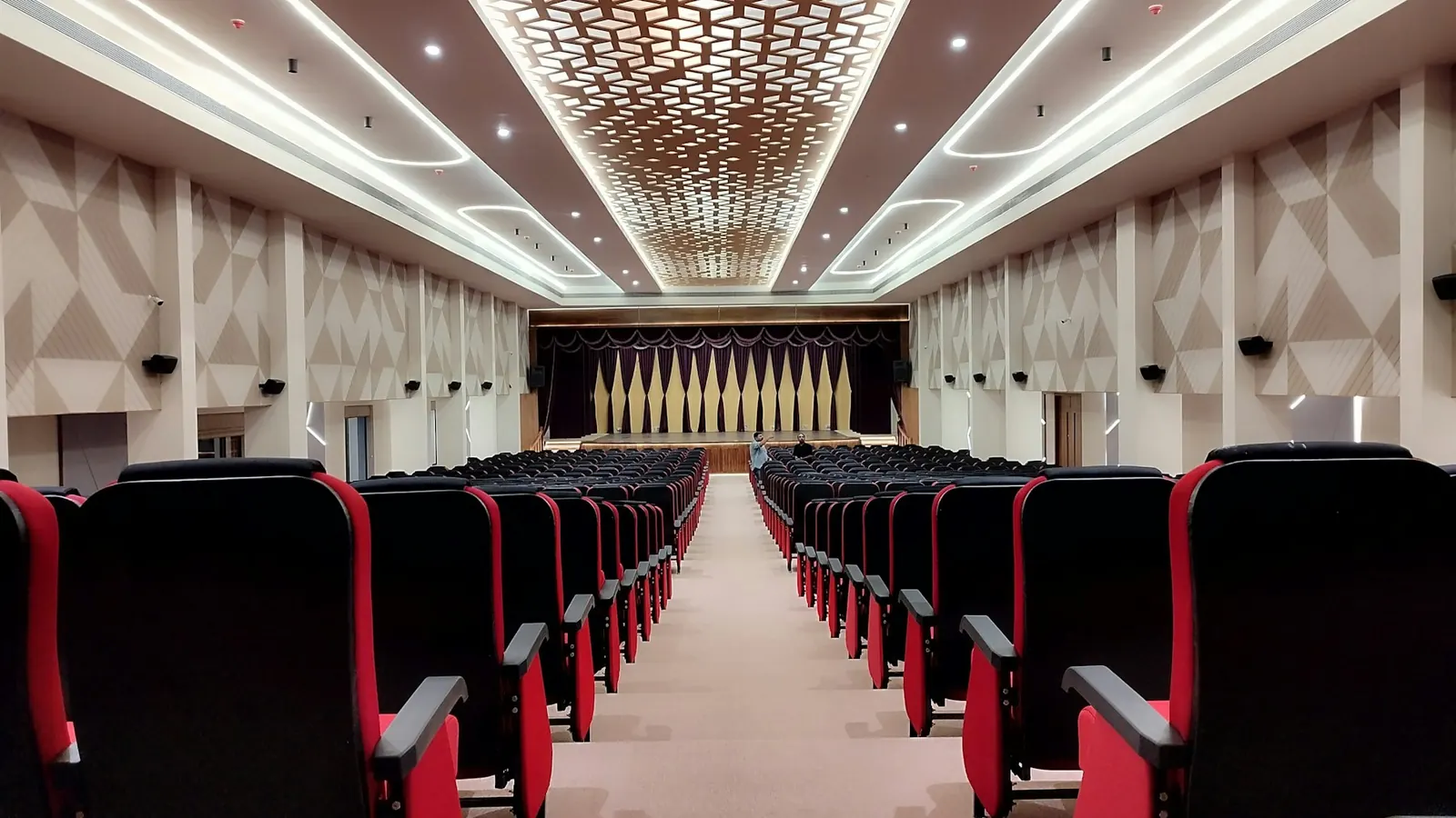 Empty auditorium with rows of red seats facing a stage under a patterned ceiling