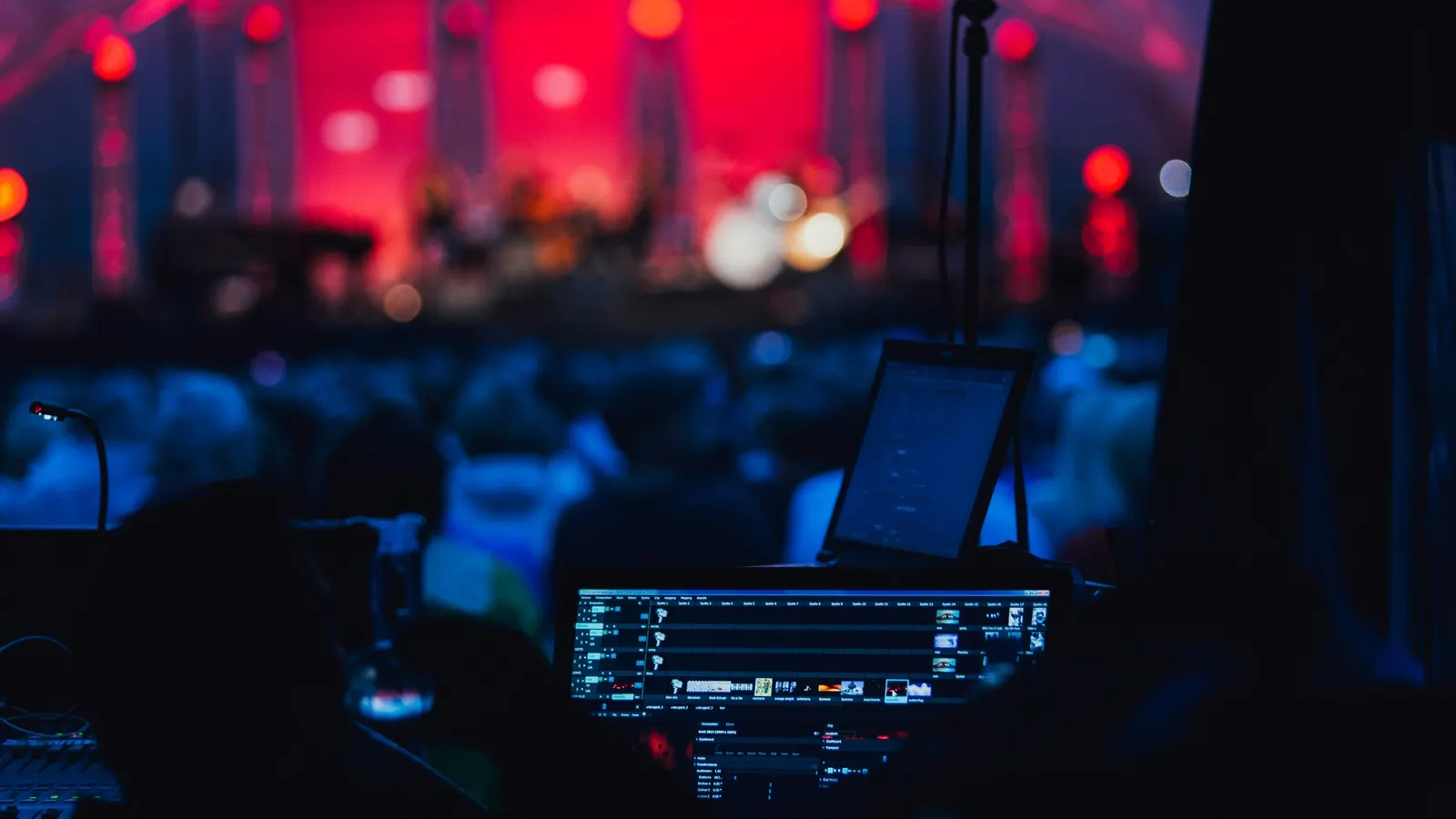 A production desk with a screen and a microphone, facing a lit stage and a seated audience at a live concert