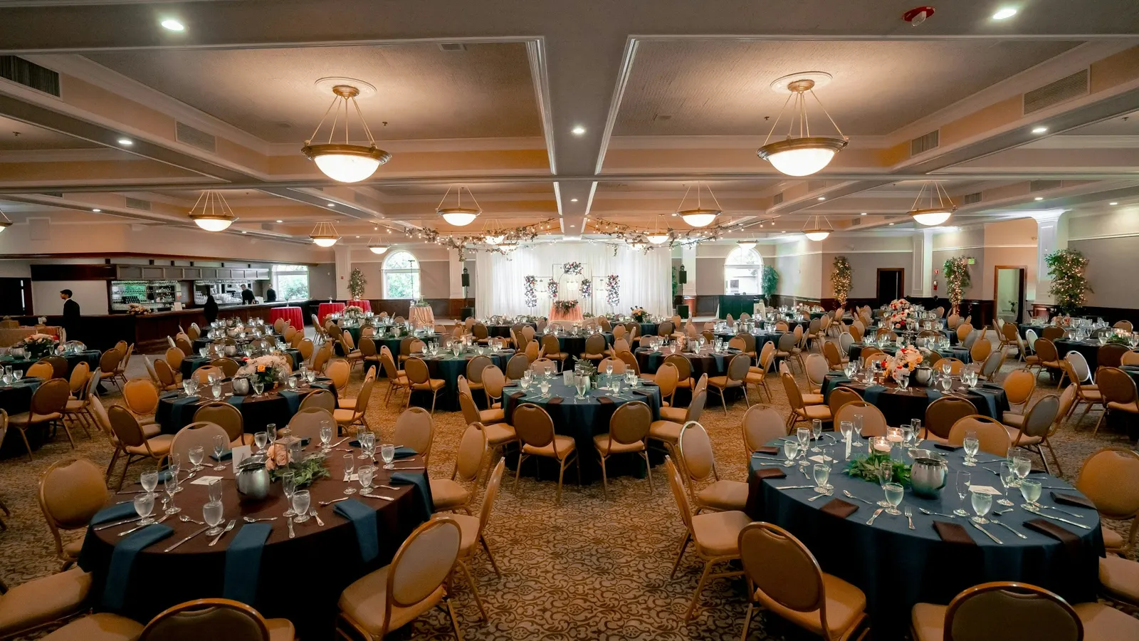 A large banquet room set for an event, with round tables, dark tablecloths, glasses and chairs under pendant lights