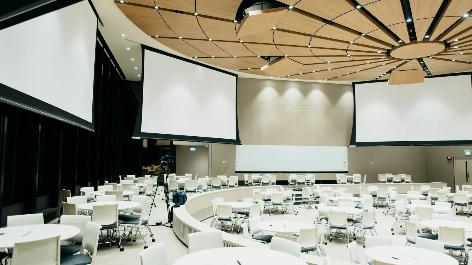 An empty conference hall with round tables, white chairs and three projection screens before an event