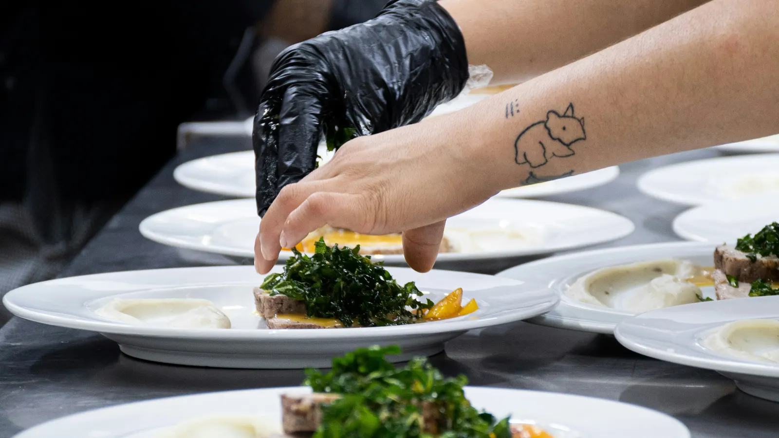 A gloved hand placing greens on a row of white plates on a steel counter in a professional kitchen