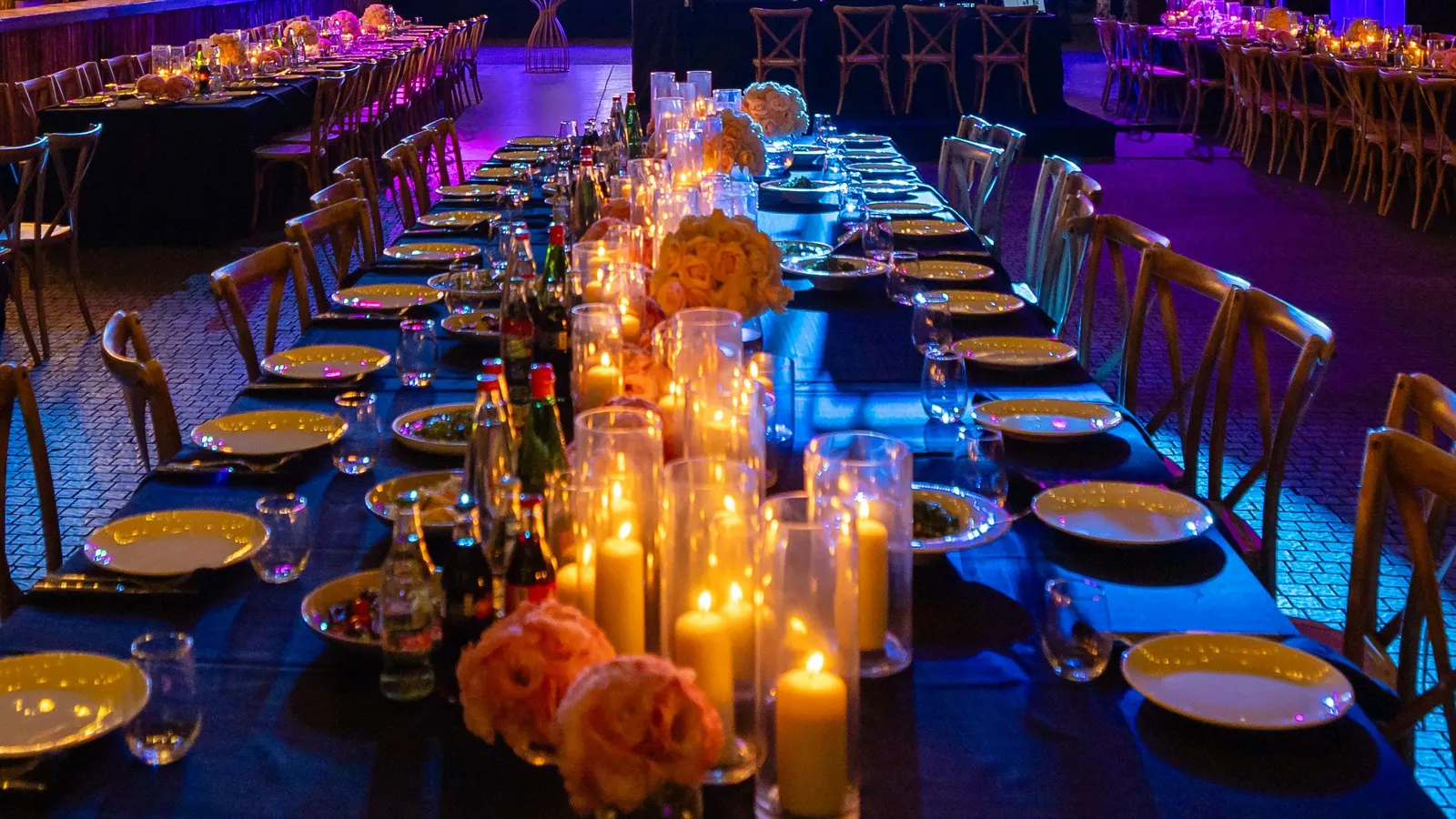 Long dinner tables set with plates, glasses and candles in tall holders, under blue and purple lights in a dark event hall