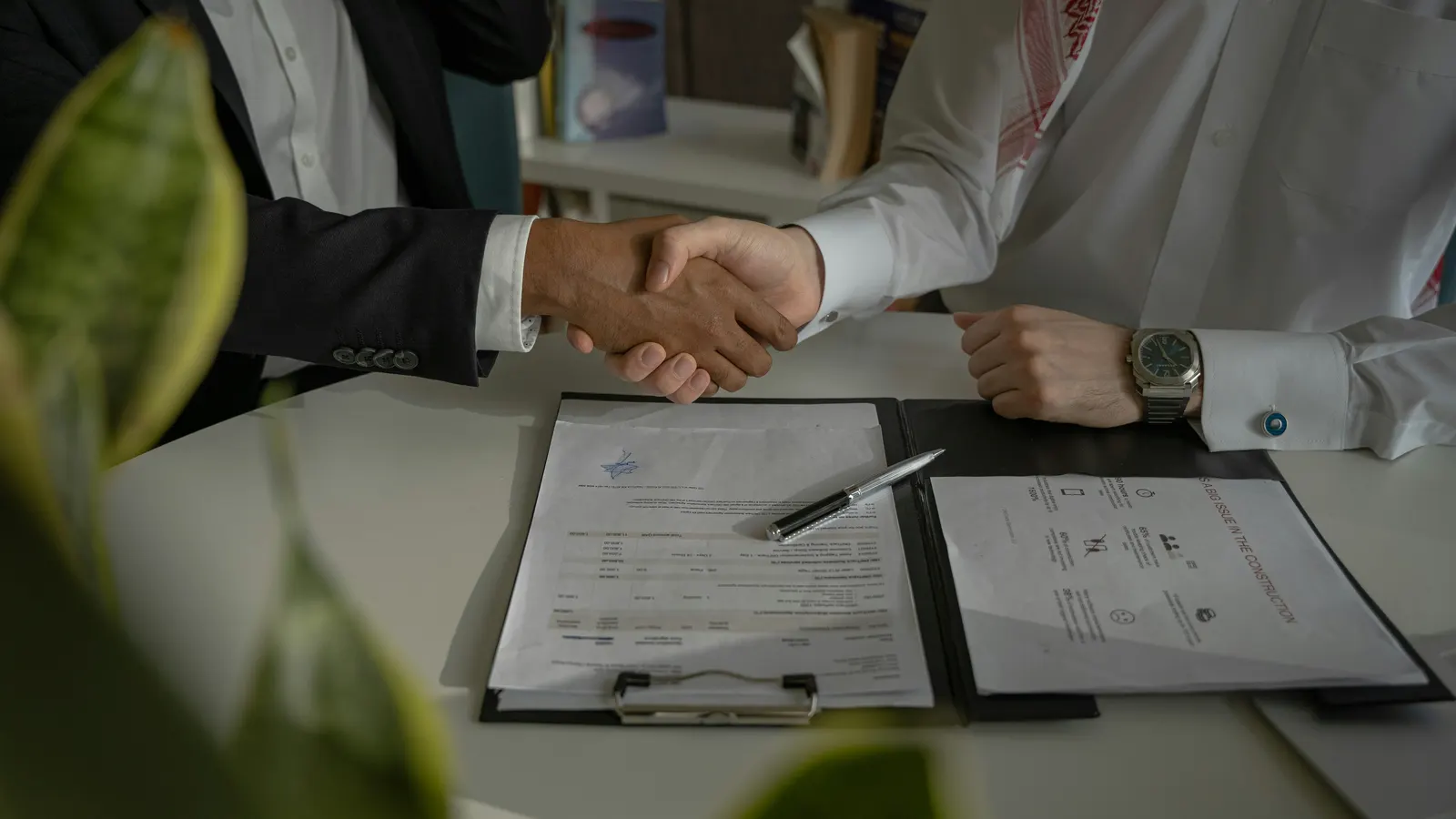 Two people shaking hands across a table, with a contract on a clipboard and a pen in front of them